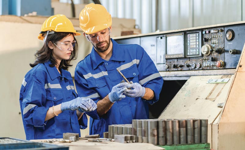 Two factory workers in yellow hard hats and blue jumpsuits inspecting metal parts at a machine.