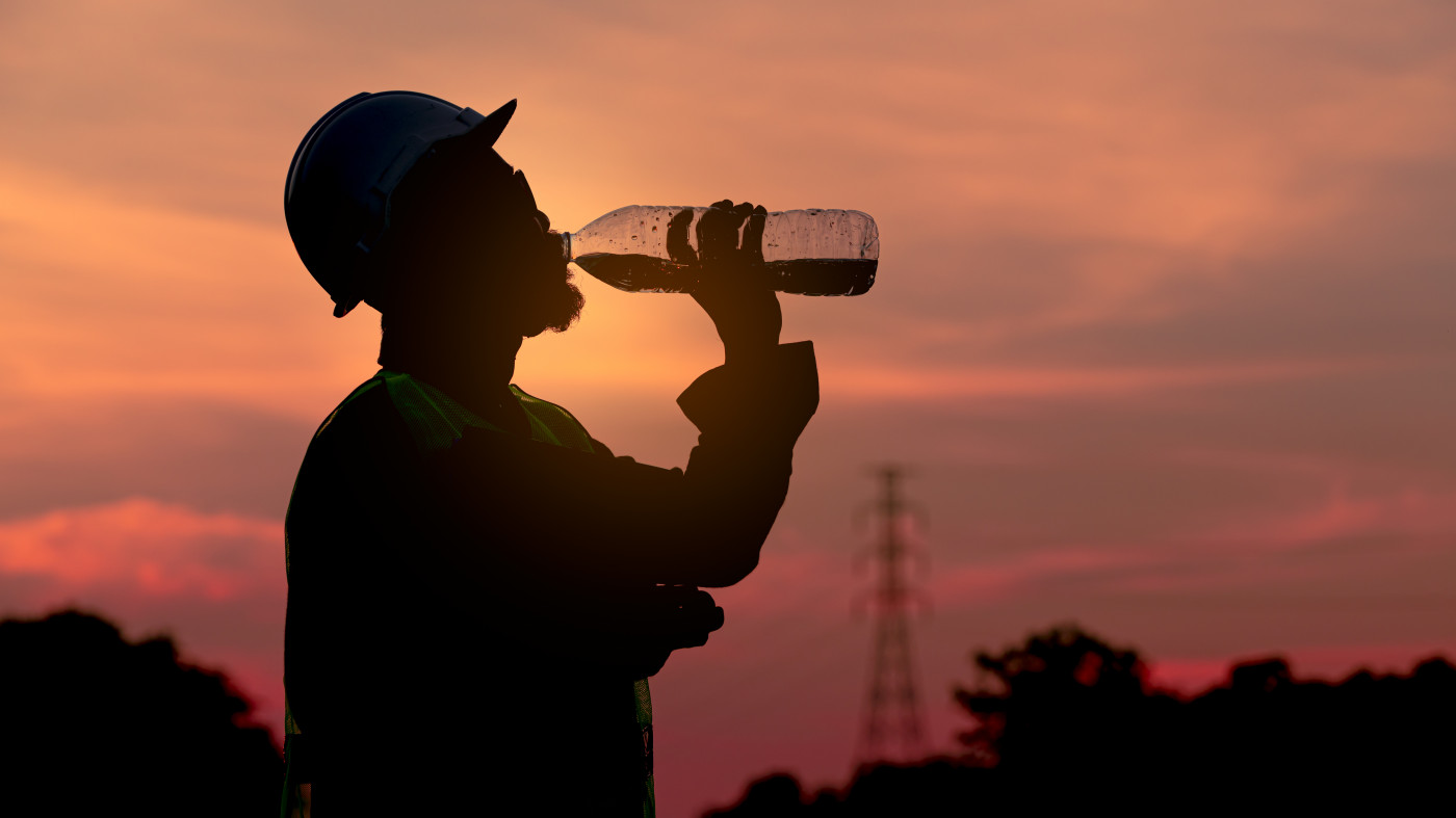 Silhouette of a construction worker wearing a hard hat drinking water from a bottle at sunset.
