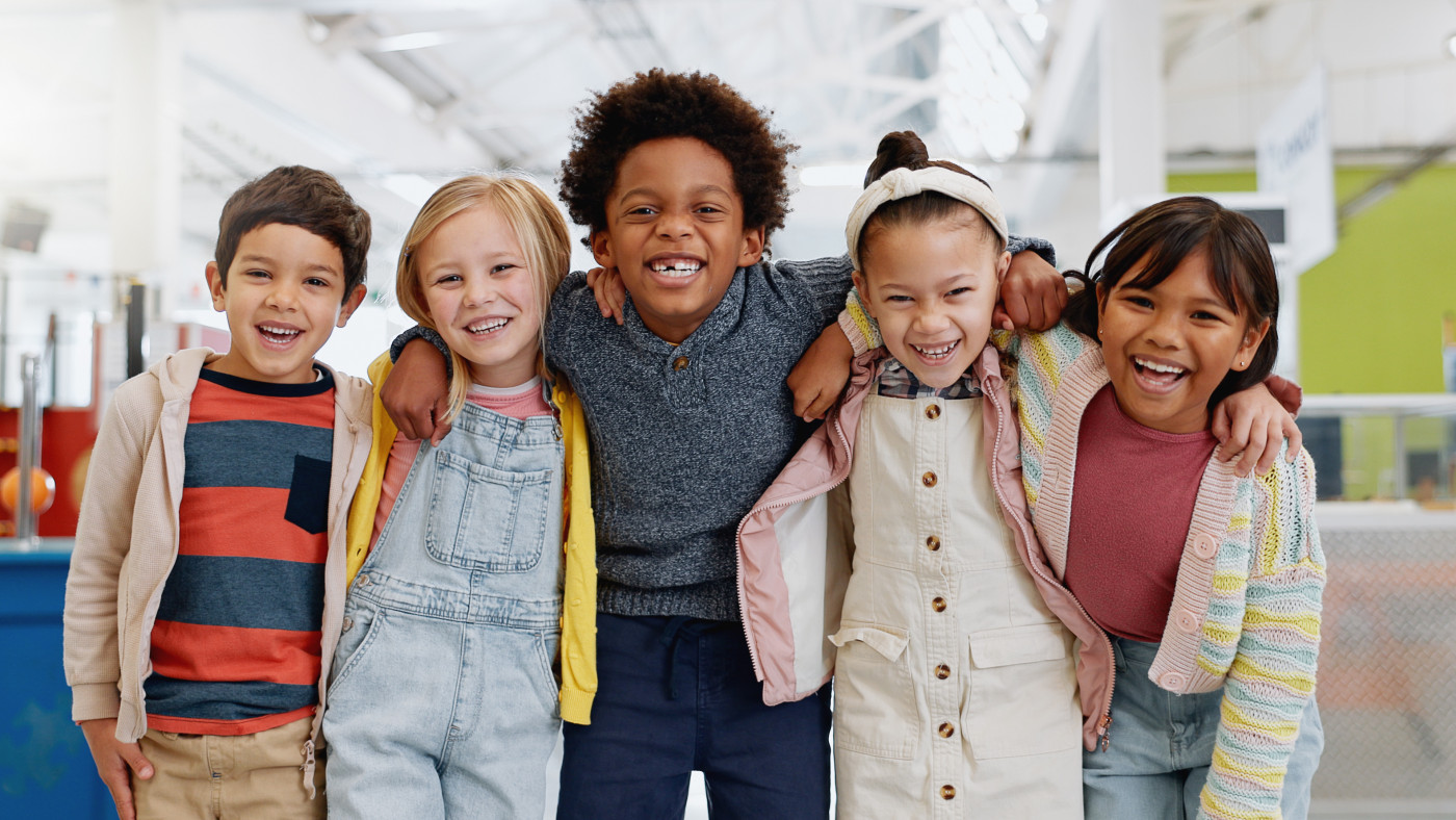 Group of five smiling children standing close together with their arms around each other in a bright indoor setting.