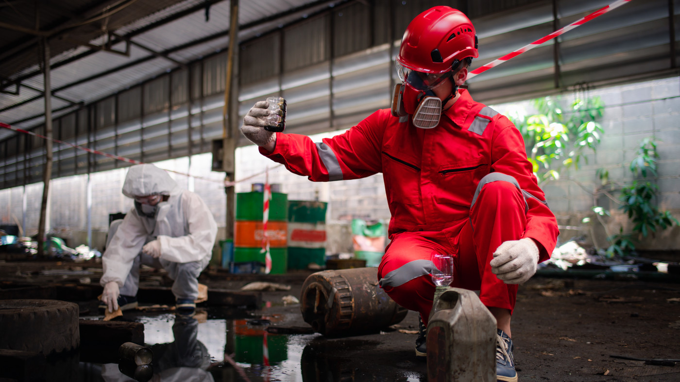 Hazmat worker in red protective gear kneels in an industrial area, holding a sample container while another worker in white protective clothing cleans up a chemical spill nearby.