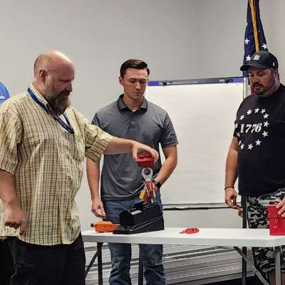 Five people watch as a participant demonstrates lockout/tagout safety equipment on a table during a classroom training session.